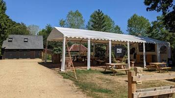 Un espace de restauration extérieur rustique, doté d’un pavillon couvert, de tables en bois et d’un chemin de gravier menant à une structure ressemblant à une grange.