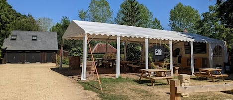 A rustic outdoor dining area with a covered pavilion, wooden tables, and a gravel path leading to a barn-like structure.