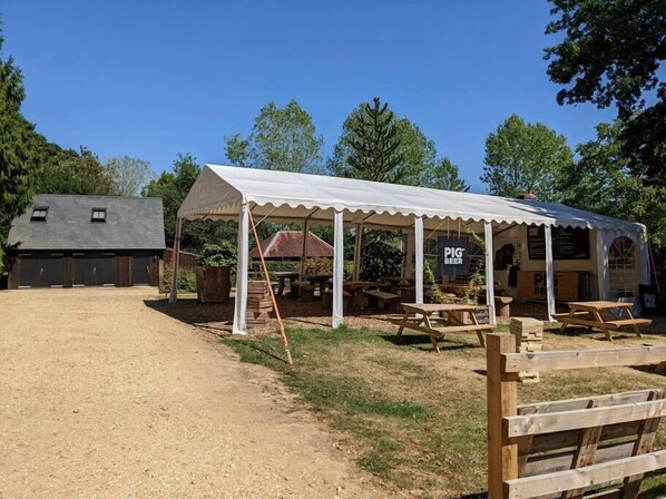 A rustic outdoor dining area with a covered pavilion, wooden tables, and a gravel path leading to a barn-like structure. - Cabin in the Forest. (Brockenhurst)