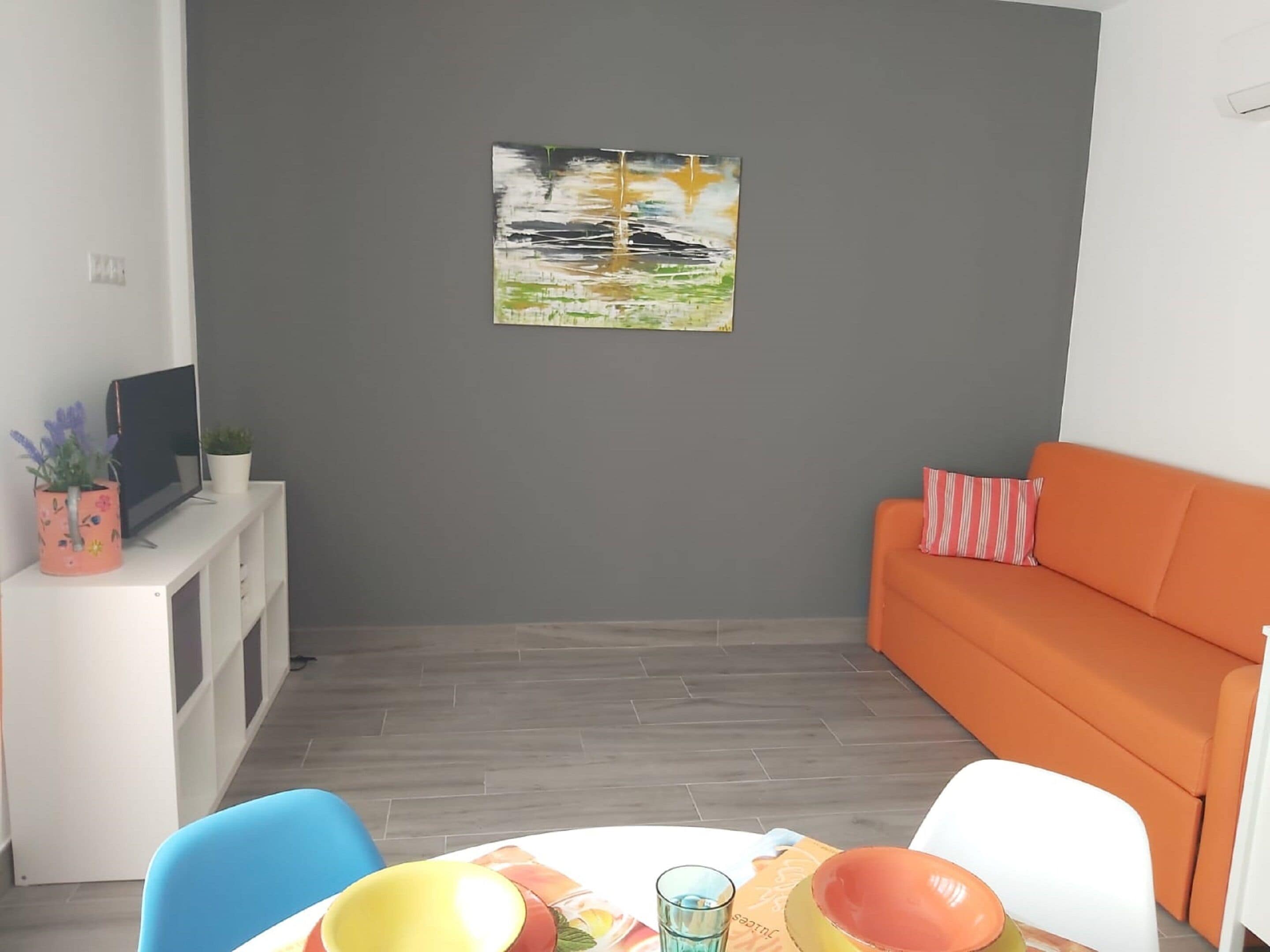 Una sala de estar moderna con una pared gris, un cuadro, un sofá naranja, un sofá blanco y una mesa de comedor con sillas.
