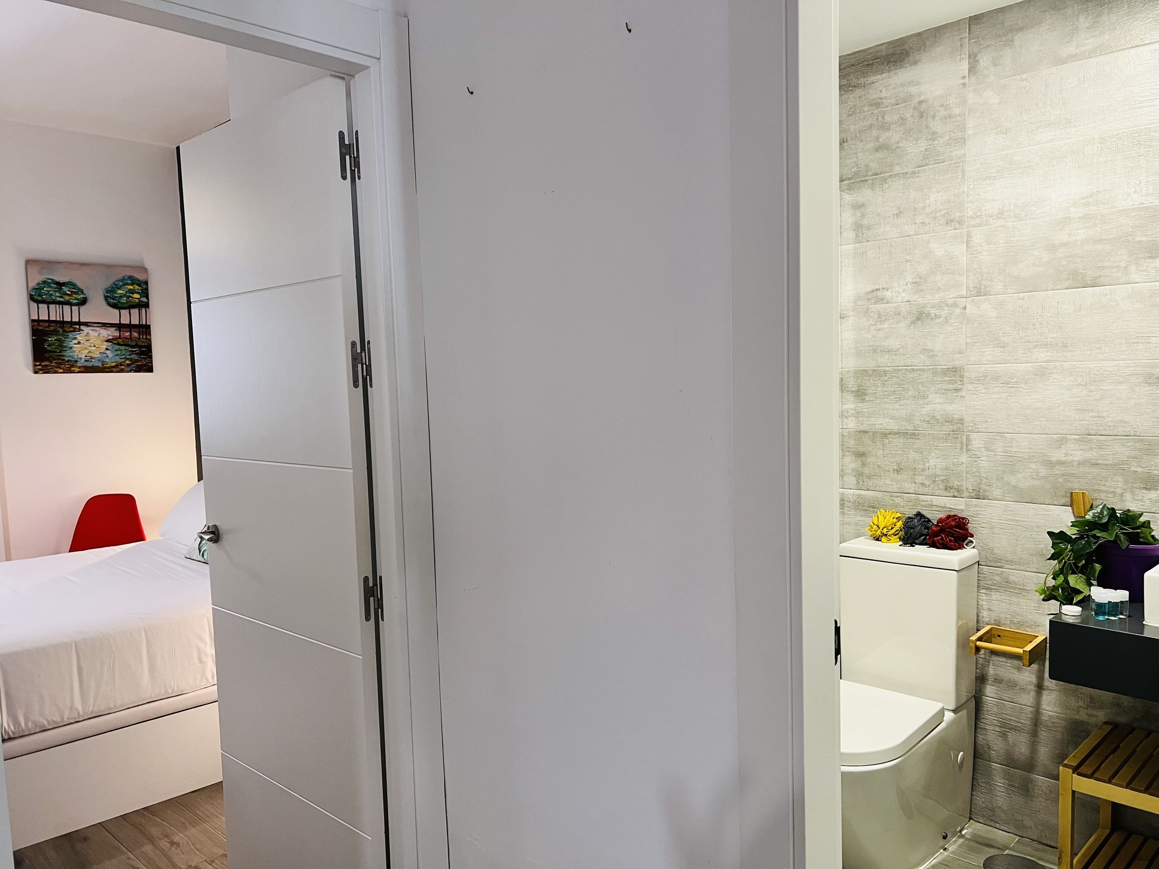 Una habitación de hotel con cama, una silla roja, un armario blanco y un baño con inodoro, lavabo y una pequeña mesa de madera.