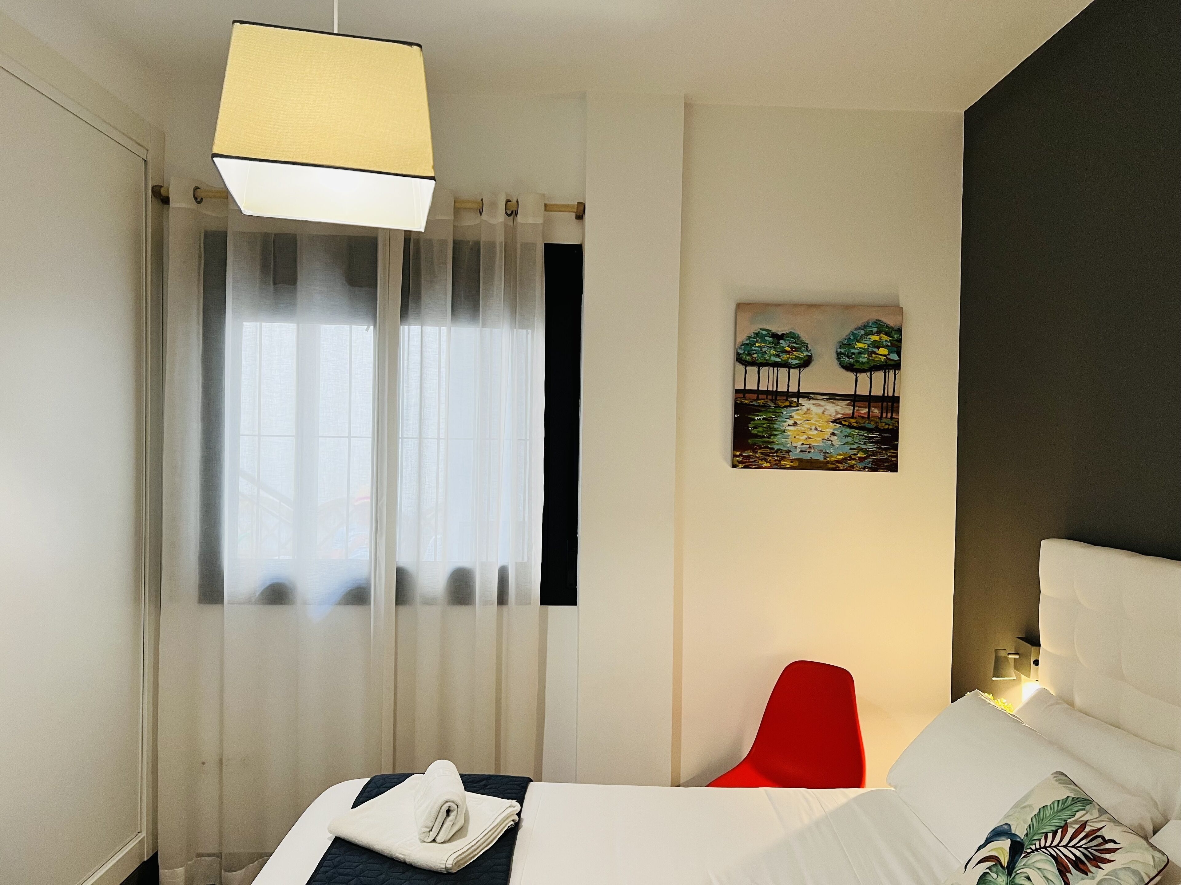 A hotel room with a bed, a red chair, a painting, and a window with curtains.