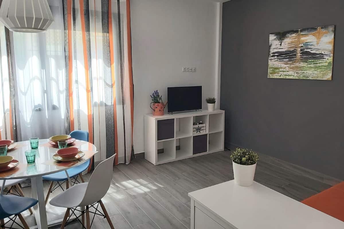 A modern living room with a dining area, a flat-screen TV on a white stand, and a painting on the wall.