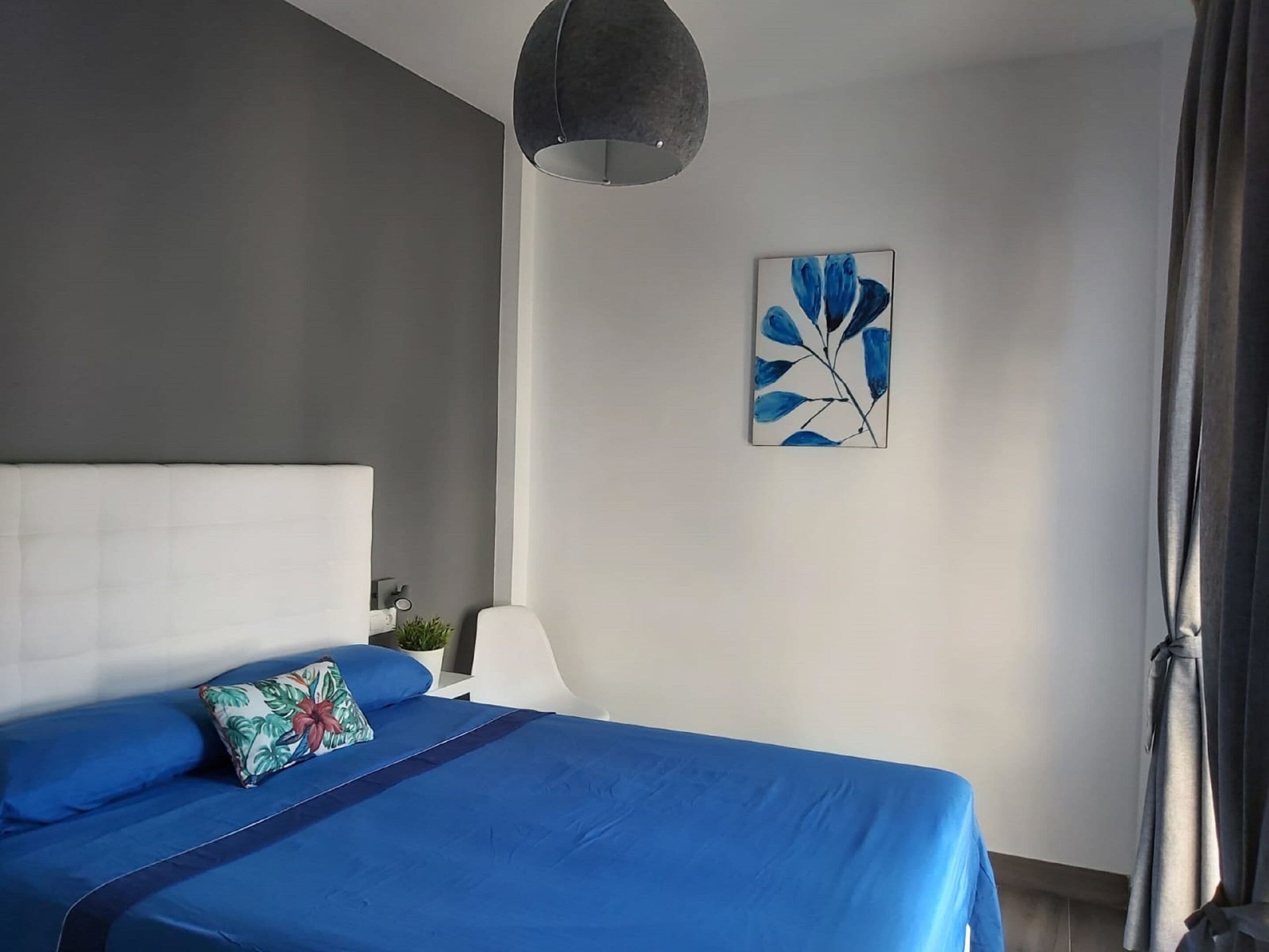 Une chambre avec un grand lit, une tête de lit moderne, un luminaire au plafond, un tableau accroché au mur et une fenêtre avec des rideaux.