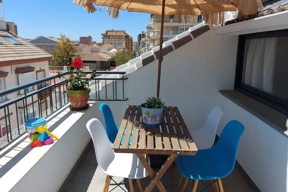 A balcony with a wooden table and blue chairs, a potted plant, and a straw umbrella.