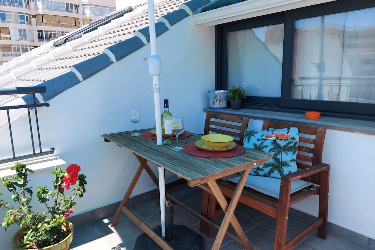 A balcony with a wooden table, two chairs, a potted plant, a wine bottle, and a window with a view of buildings.