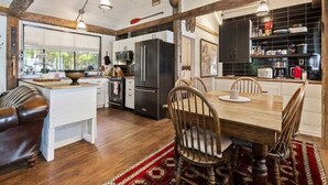 A rustic kitchen with wooden beams, a dining table with chairs, a refrigerator, and a coffee machine.