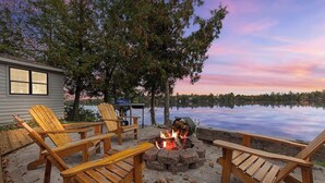 A lakeside deck with wooden chairs, a fire pit, and a view of a calm lake at sunset.