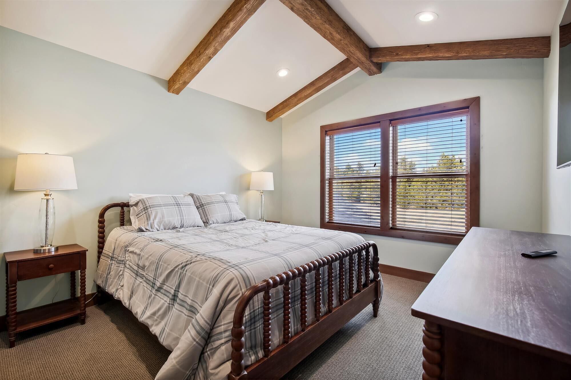 A bedroom with a wooden bed, two bedside tables, a desk, and a window with blinds.