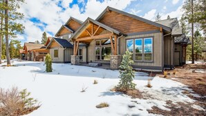 Una cabaña de troncos moderna con un porche amplio y numerosas ventanas, rodeada de nieve y árboles.