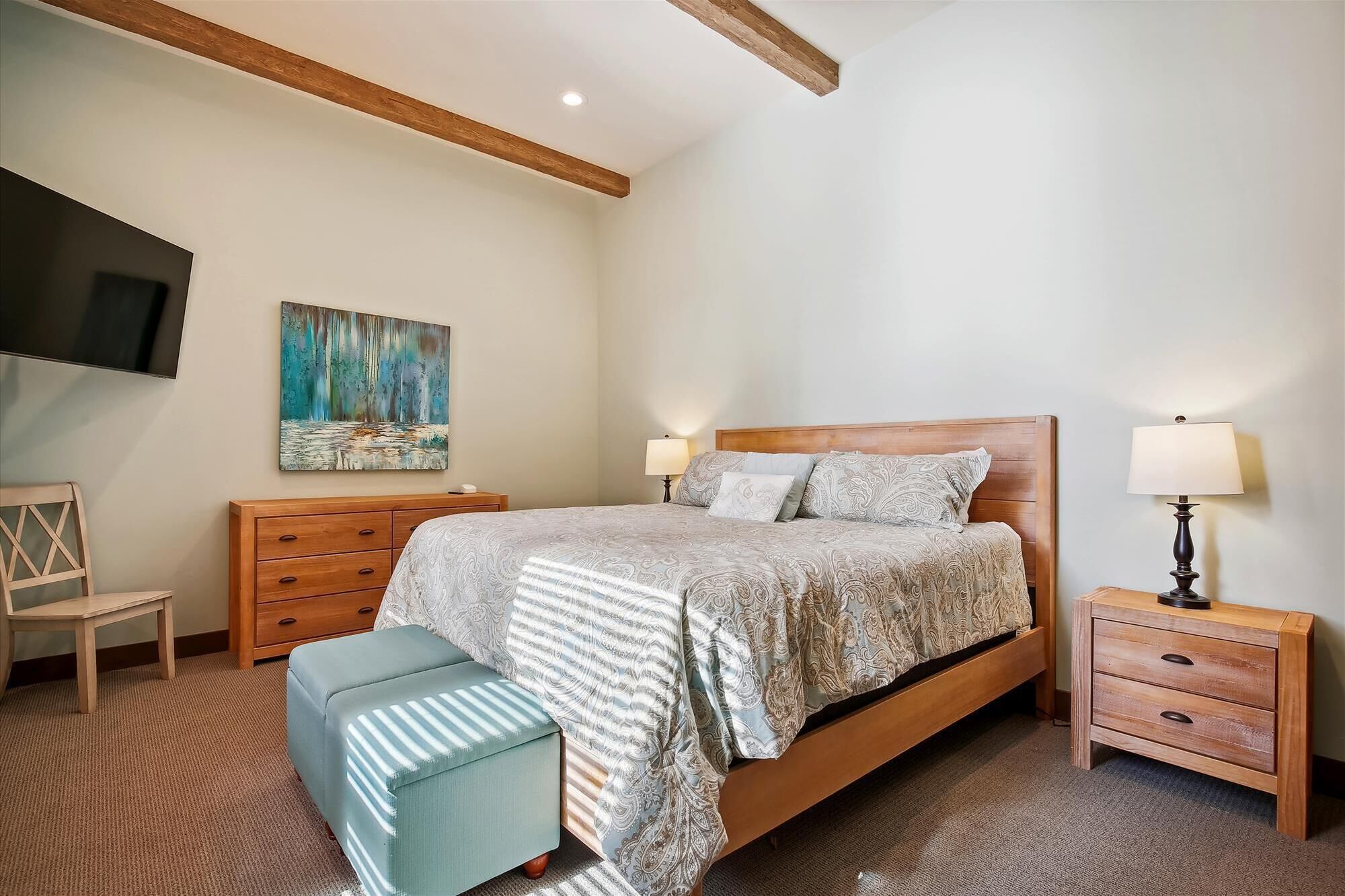 A bedroom with a wooden bed, two bedside tables, a TV mounted on the wall, and a painting above the bed.
