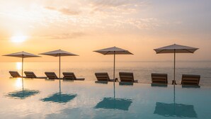 Una piscina infinita con sillones de playa y sombrillas, con vistas al mar al atardecer.