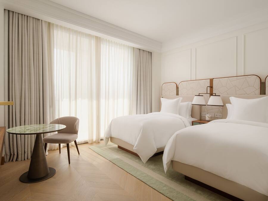 Quarto de hotel com duas camas, uma mesa redonda, uma cadeira e um criado-mudo com luminárias.