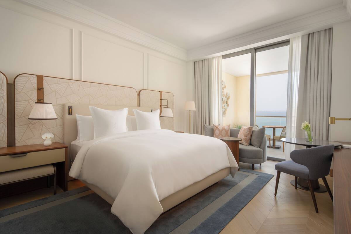Une chambre d’hôtel avec un grand lit, deux tables de chevet avec des lampes, une petite table avec une chaise, et une vue sur l’extérieur grâce à une grande fenêtre.