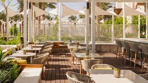 Een terras buiten met tafels en stoelen, beschaduwd door witte parasols, omgeven door palmbomen en een sierhekwerk.