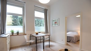 Ein heller, moderner Raum mit Holzboden, einem Esstisch mit Stühlen, einer kleinen weißen Kommode und einem Bett im angrenzenden Zimmer.