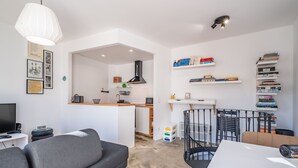 A modern living room with a grey sofa, a white table, and a red rug. There is a kitchen area with a white countertop and a black stove hood. Bookshelves are mounted on the wall, and a black metal chair is visible. - Casa Luar - By Wehost (Funchal)