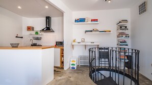 A compact kitchen with a white countertop, a black range hood, and a microwave. There are white shelves with books and a black spiral staircase. - Casa Luar - By Wehost (Funchal)
