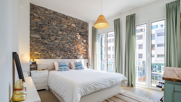 A bedroom with a stone wall, a bed with white bedding, a nightstand, a desk with a lamp, and a view of buildings through large windows. - Casa Luar - By Wehost (Funchal)