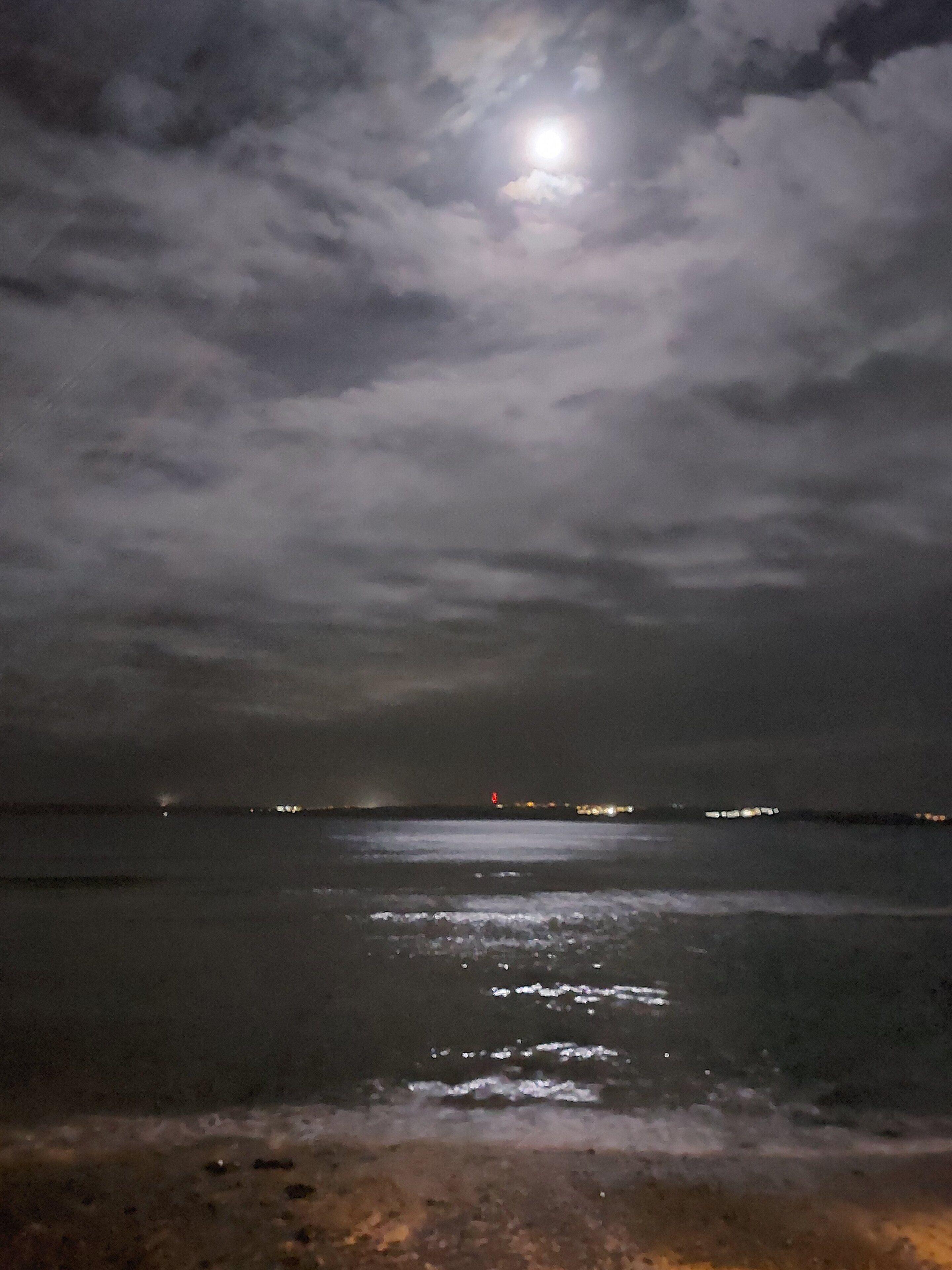 Une vue nocturne d’une plage, avec un ciel nuageux et une lune visible.