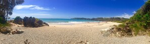 A pebbly beach with rocky outcrops, clear blue water, and a distant forested hill. - “The Cove” 
Beachfront Retreat.
Wake to the Ocean. Unwind in Nature. (Korora)