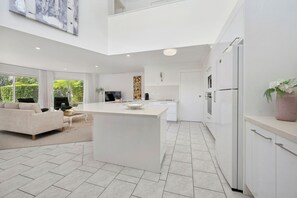A modern kitchen with a central island, white cabinetry, and a dining area with a sofa and coffee table. - “The Cove” 
Beachfront Retreat.
Wake to the Ocean. Unwind in Nature. (Korora)