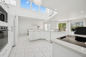 A modern kitchen with white cabinetry, a central island, and a large window providing natural light. - “The Cove” 
Beachfront Retreat.
Wake to the Ocean. Unwind in Nature. (Korora)