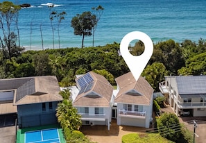 A residential area with houses featuring sloped roofs and solar panels, surrounded by lush greenery and a beach. - “The Cove” 
Beachfront Retreat.
Wake to the Ocean. Unwind in Nature. (Korora)