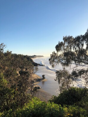 A coastal landscape with a rocky outcrop in the foreground, a sandy beach, and dense foliage.