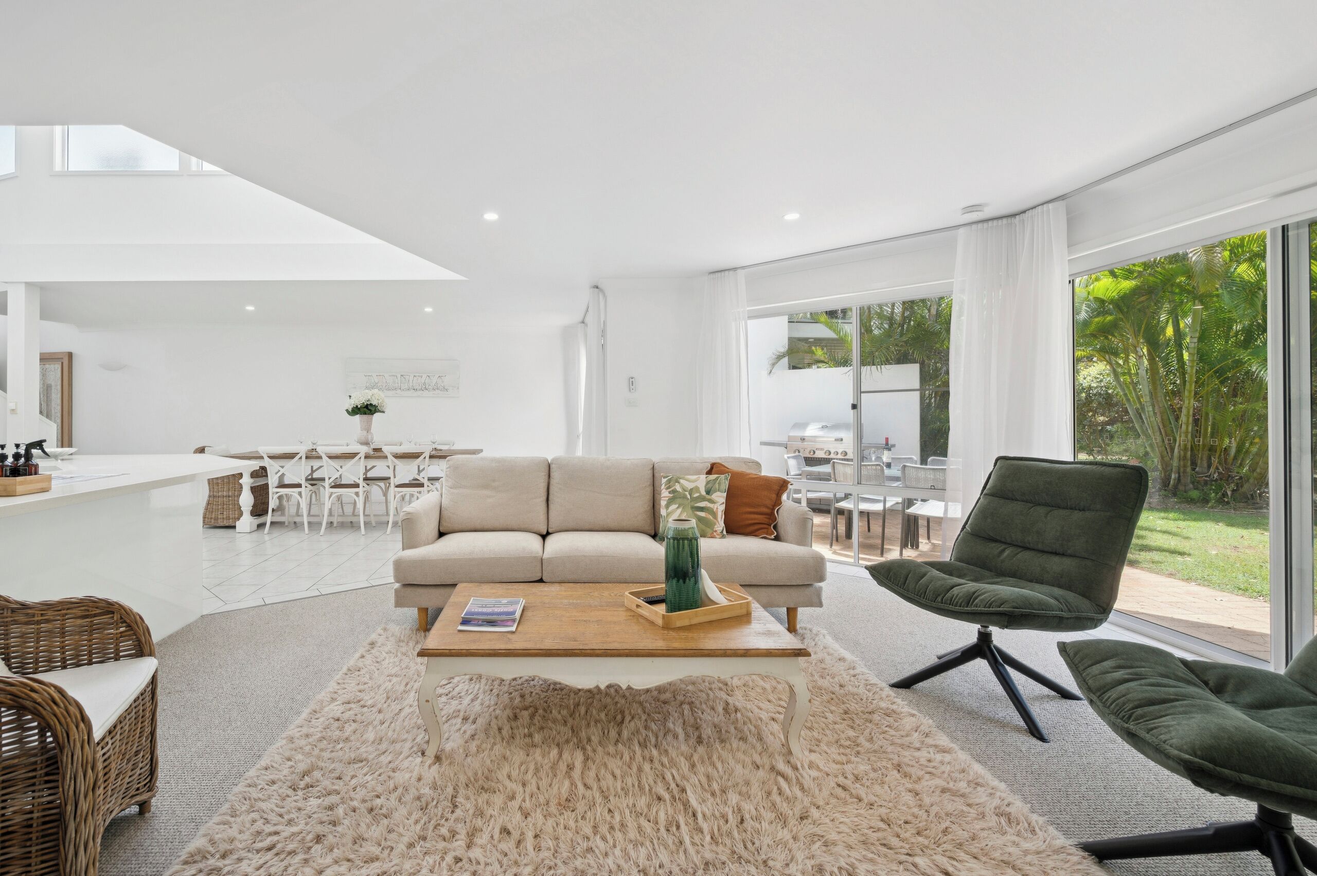 A modern living room with a sofa, coffee table, and chairs, a kitchen area in the background, and large windows with a view of greenery.