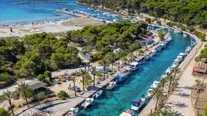 Un port de plaisance avec des bateaux amarrés le long d’une promenade, un pont piétonnier et une zone de plage où se trouvent des personnes et des parasols.