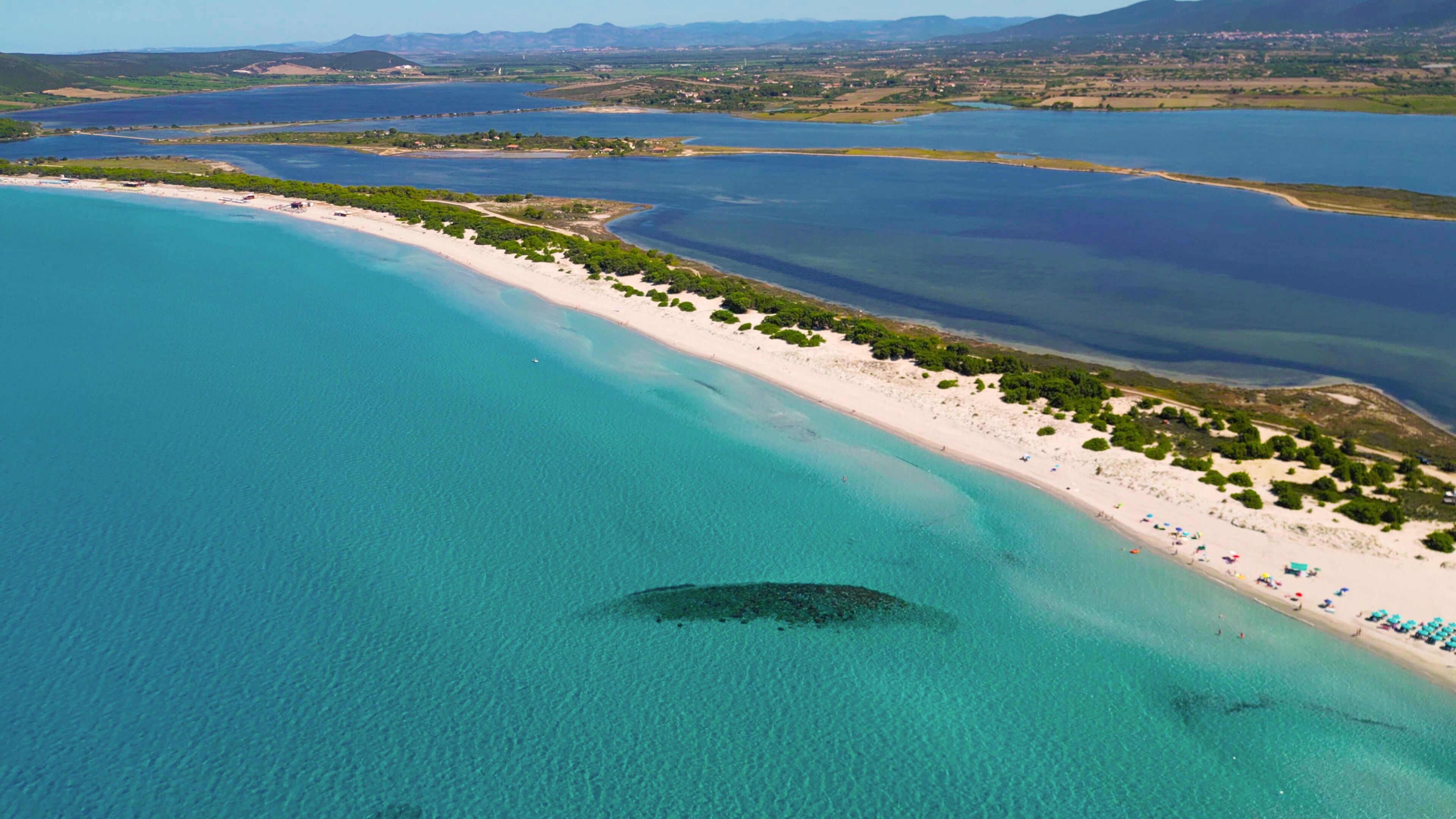 Vista aérea de una zona costera con playa arenosa, aguas turquesas cristalinas y colinas con un pueblo distante al fondo.