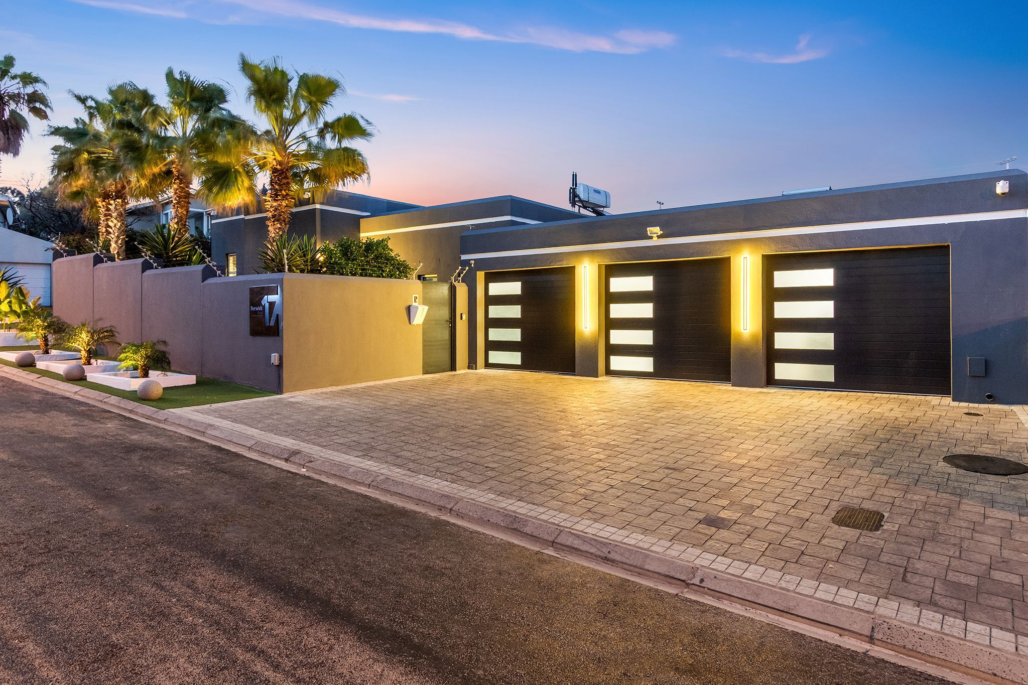 A modern building with a paved driveway, a garage with illuminated panels, and palm trees.