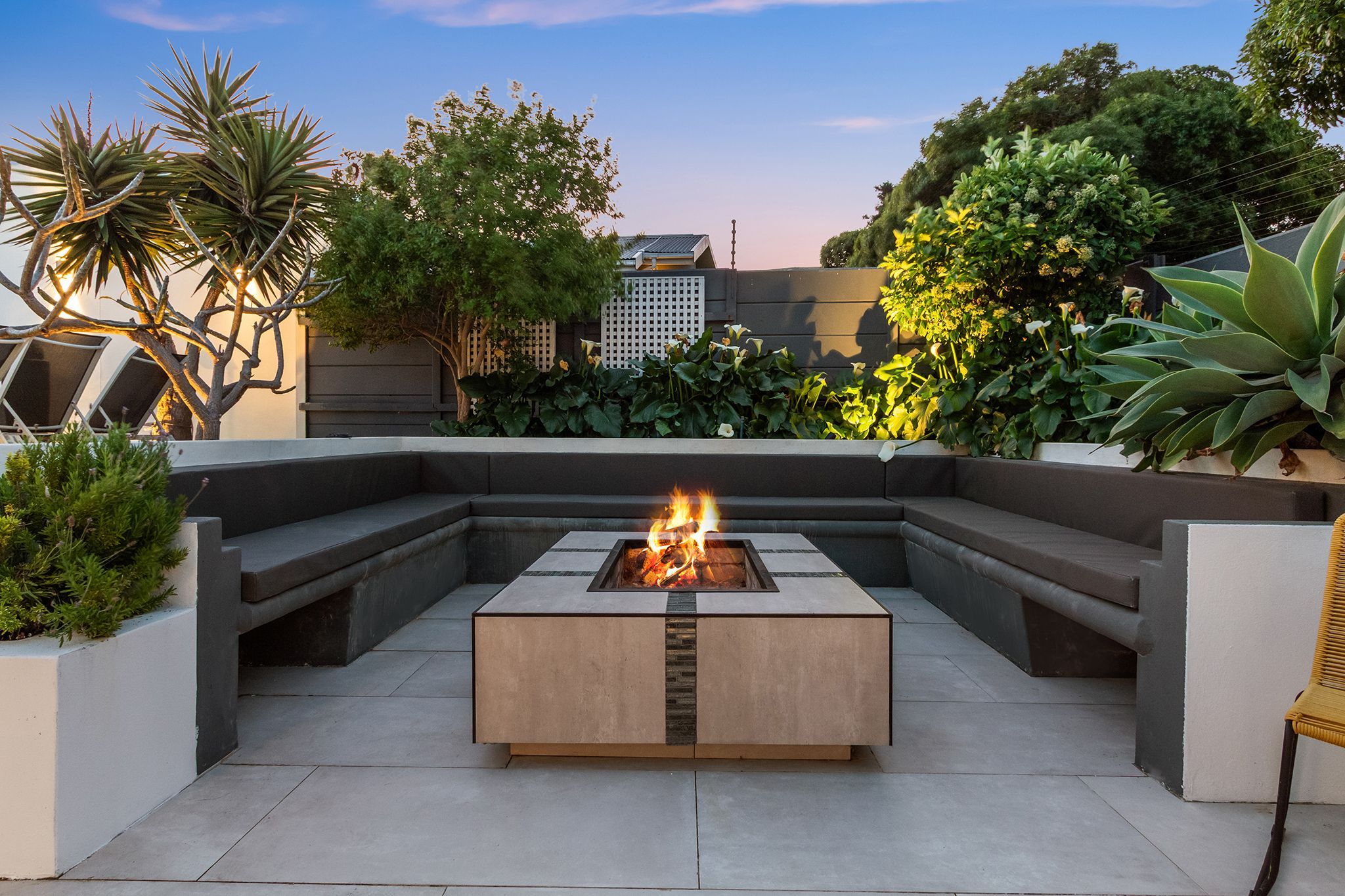 A modern outdoor seating area with a square fire pit table surrounded by built-in benches.