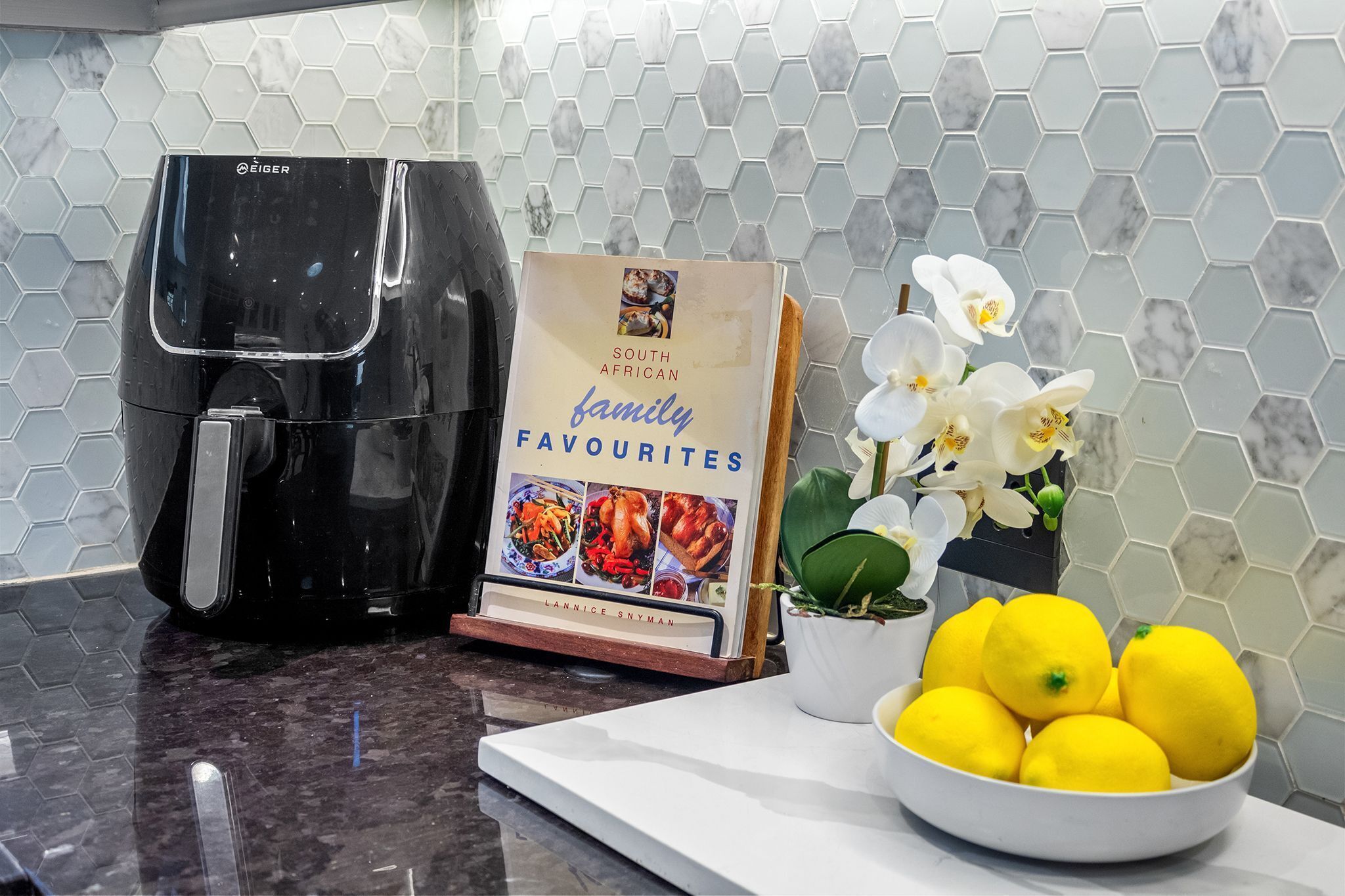 Un plan de travail de cuisine avec une machine à café, un livre de recettes intitulé « Les plats préférés des familles sud-africaines », une orchidée en pot et un bol de citrons.