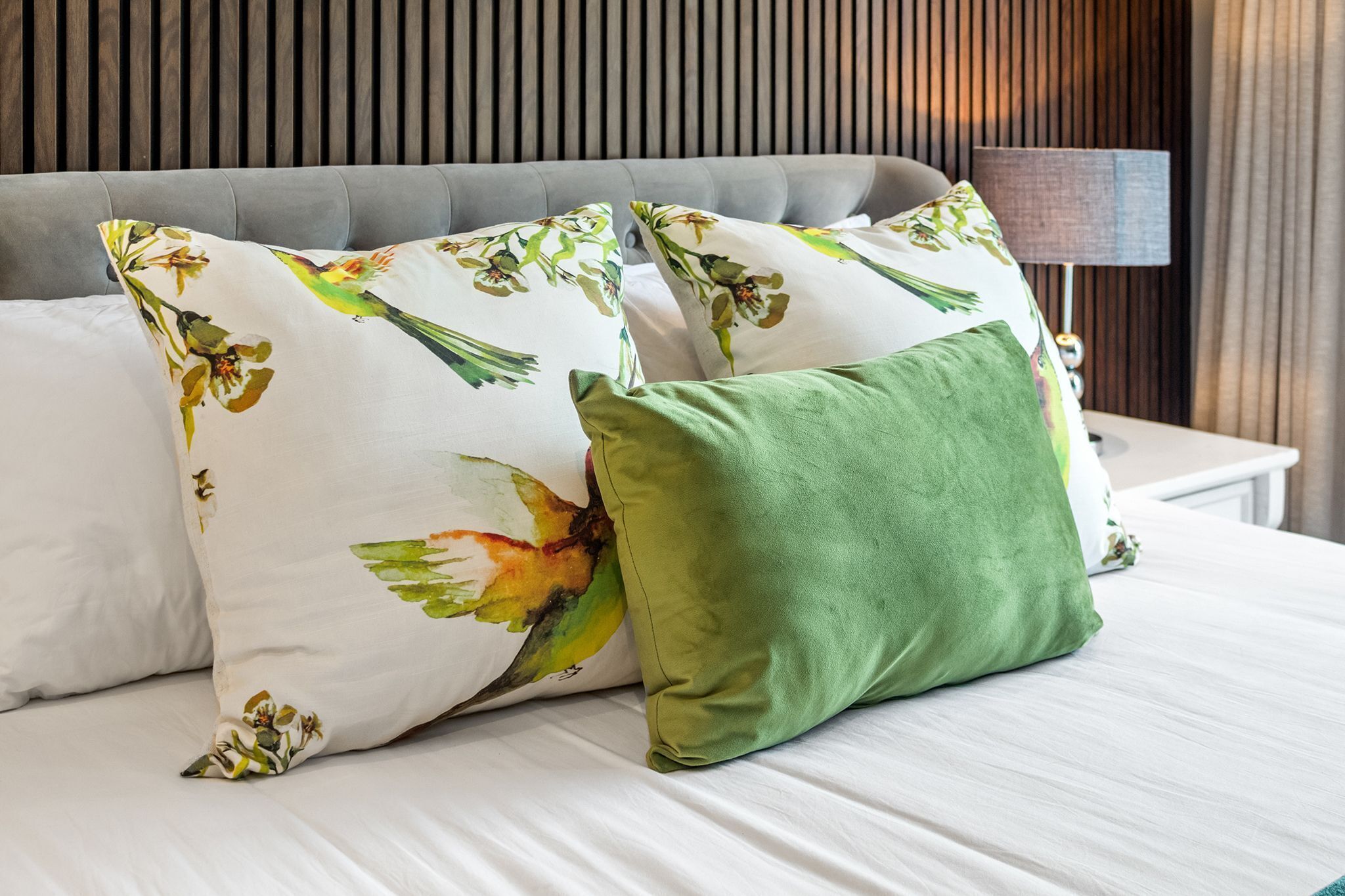 A bed with floral-patterned pillows and a green accent pillow, a grey upholstered headboard, and a bedside lamp.