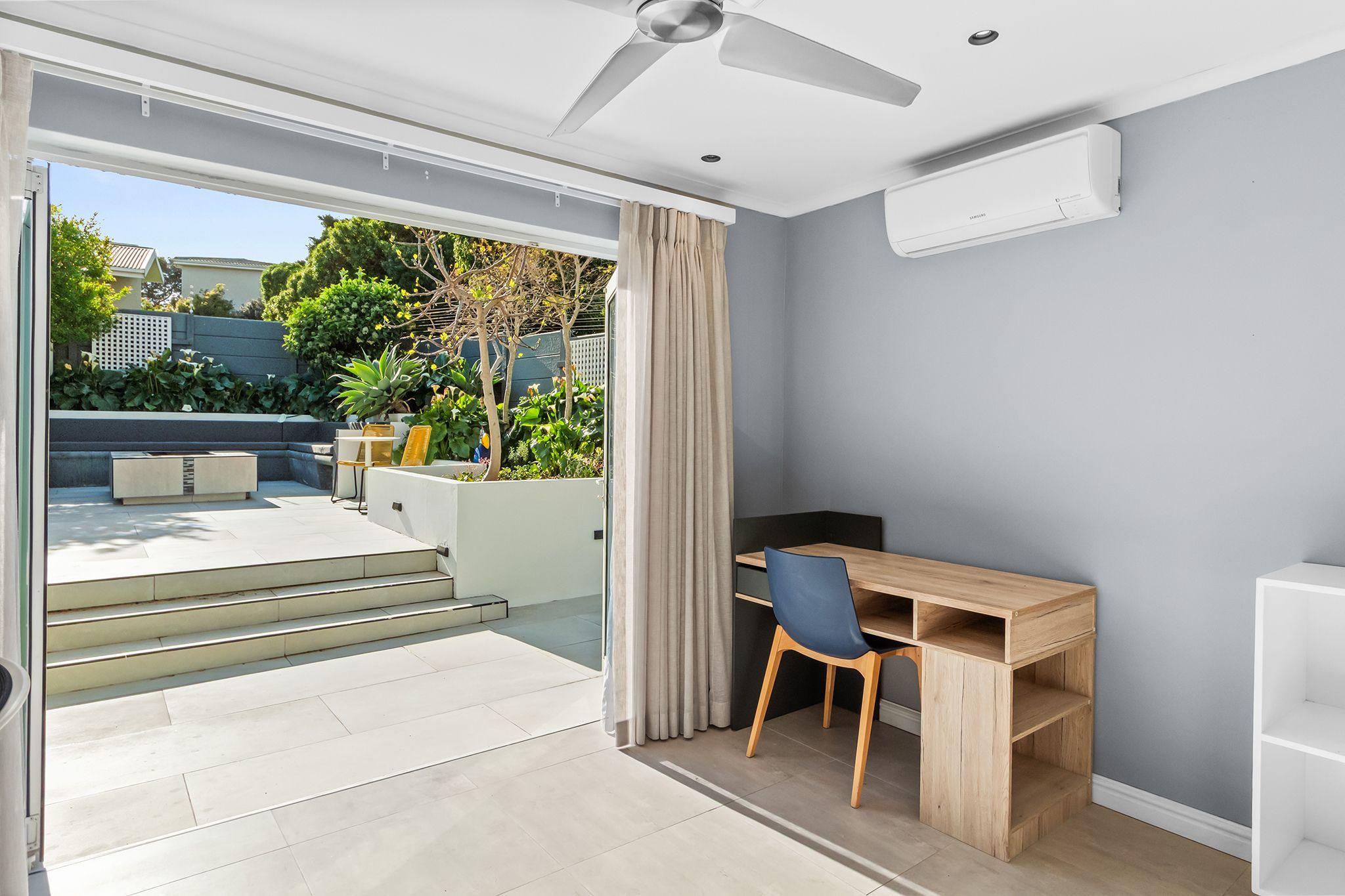 Une chambre moderne dotée d’une porte-fenêtre coulissante donnant sur un balcon, d’un bureau en bois avec une chaise et d’un ventilateur de plafond.