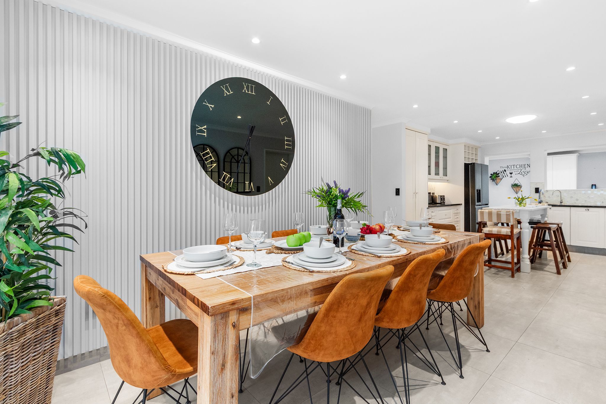 A modern dining area with a wooden table set for four, surrounded by orange chairs. A large wall clock is mounted on the wall above the table.