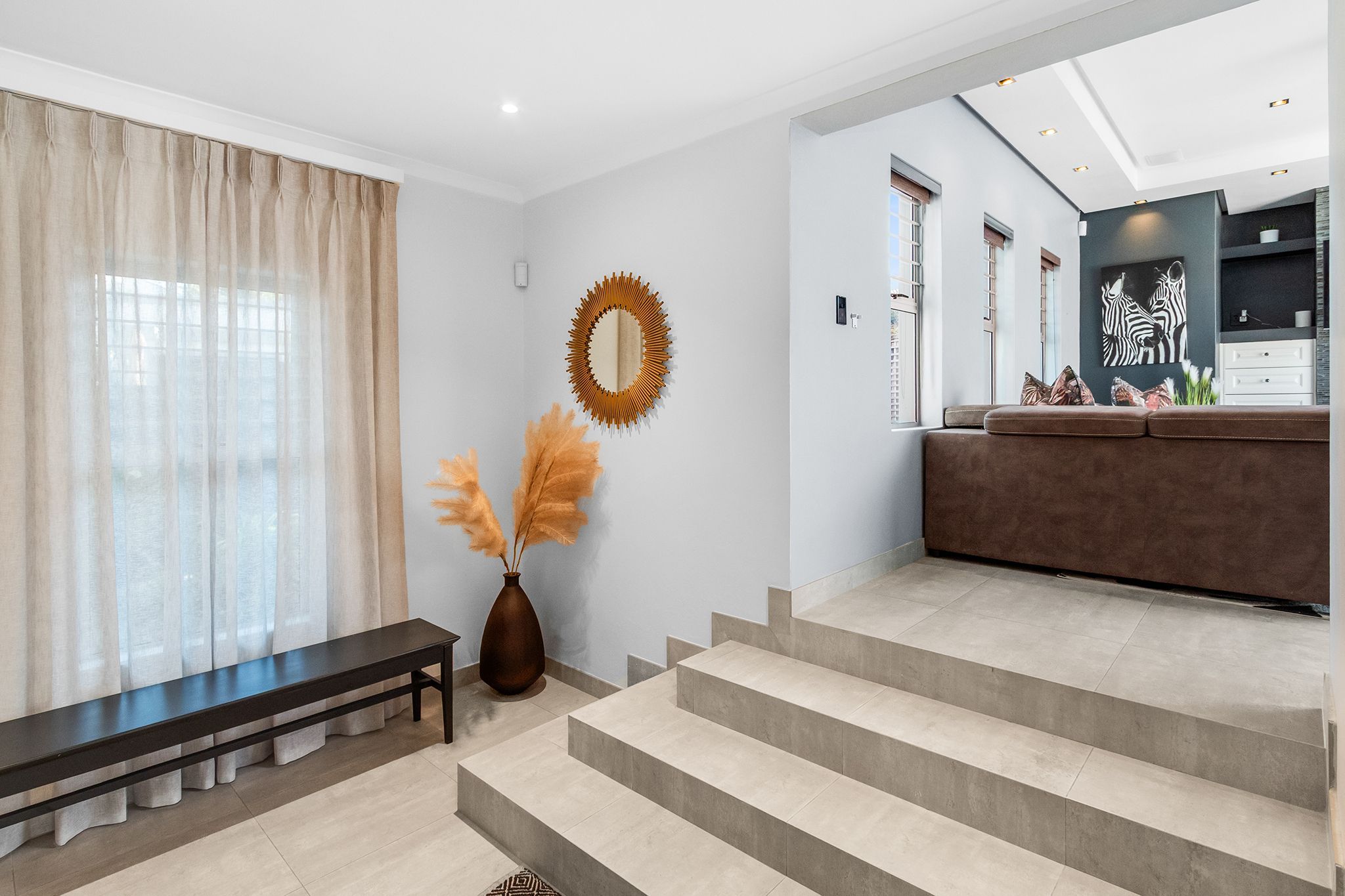 Un salon moderne avec un banc, un vase contenant de l’herbe des pampas et un escalier menant à un espace cuisine orné d’un tableau représentant un zèbre.