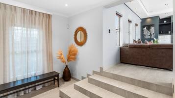 Un salon moderne avec un banc, un vase contenant de l’herbe des pampas et un escalier menant à un espace cuisine orné d’un tableau représentant un zèbre.