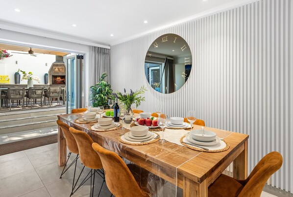 A dining area with a wooden table set for six, surrounded by orange chairs. A large mirror hangs on the wall, and a kitchen is visible in the background. - Rancho Relaxo (Cape Town)
