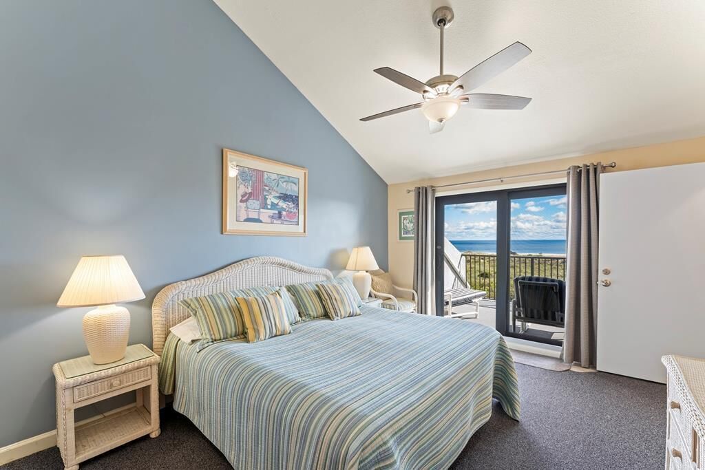 A bedroom with a bed, a ceiling fan, a nightstand with a lamp, a chair, and a balcony with a view of the ocean.