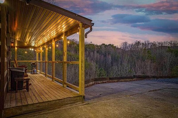 A wooden deck with string lights, a table, and chairs, overlooking a forested area.