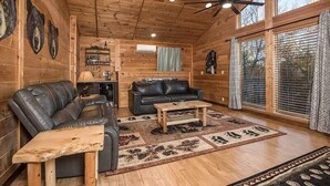 Interior de una cabaña de troncos con un sofá de cuero, una mesa de centro de madera y un ventilador de techo.