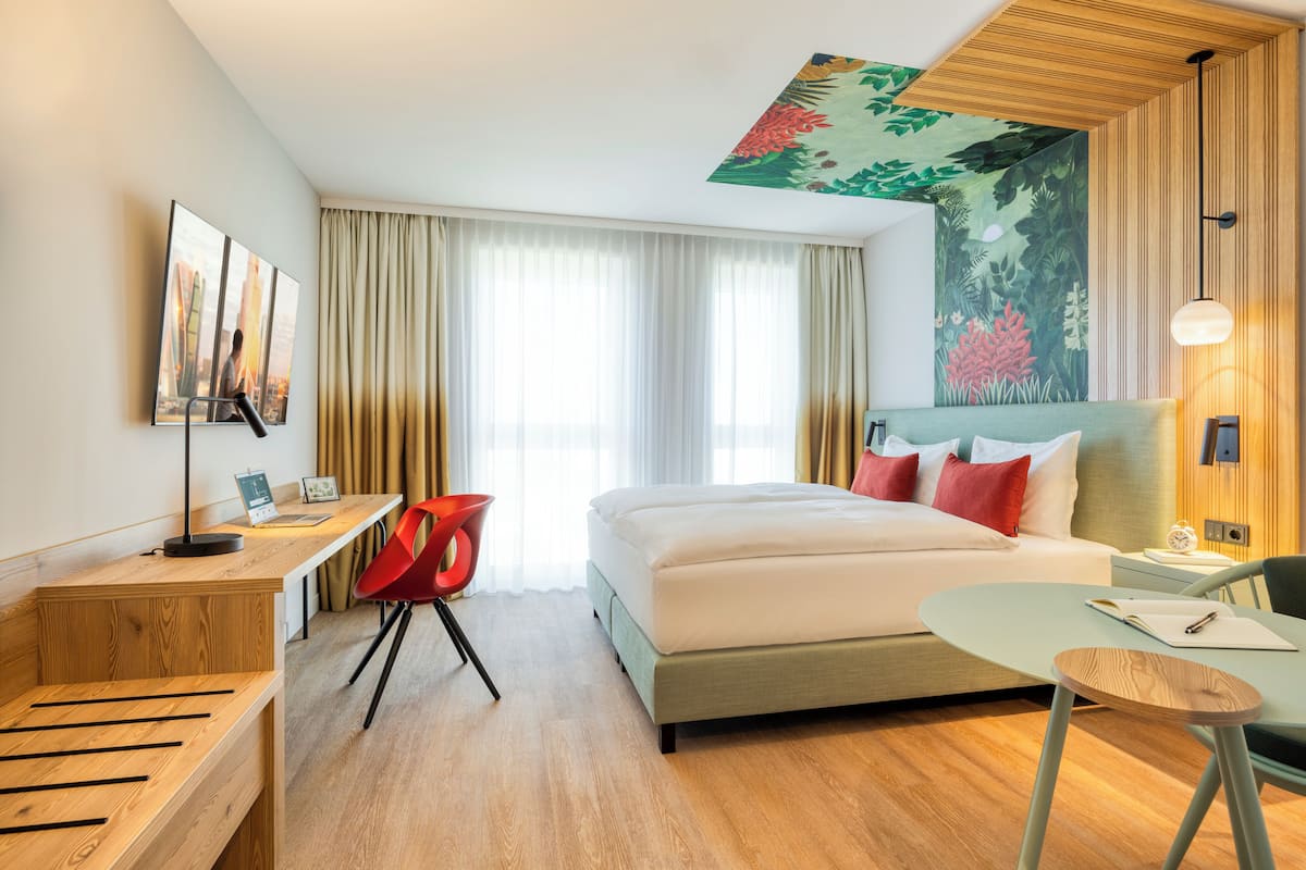 A hotel room with a bed, a desk, a chair, a small table, and a painting on the wall.