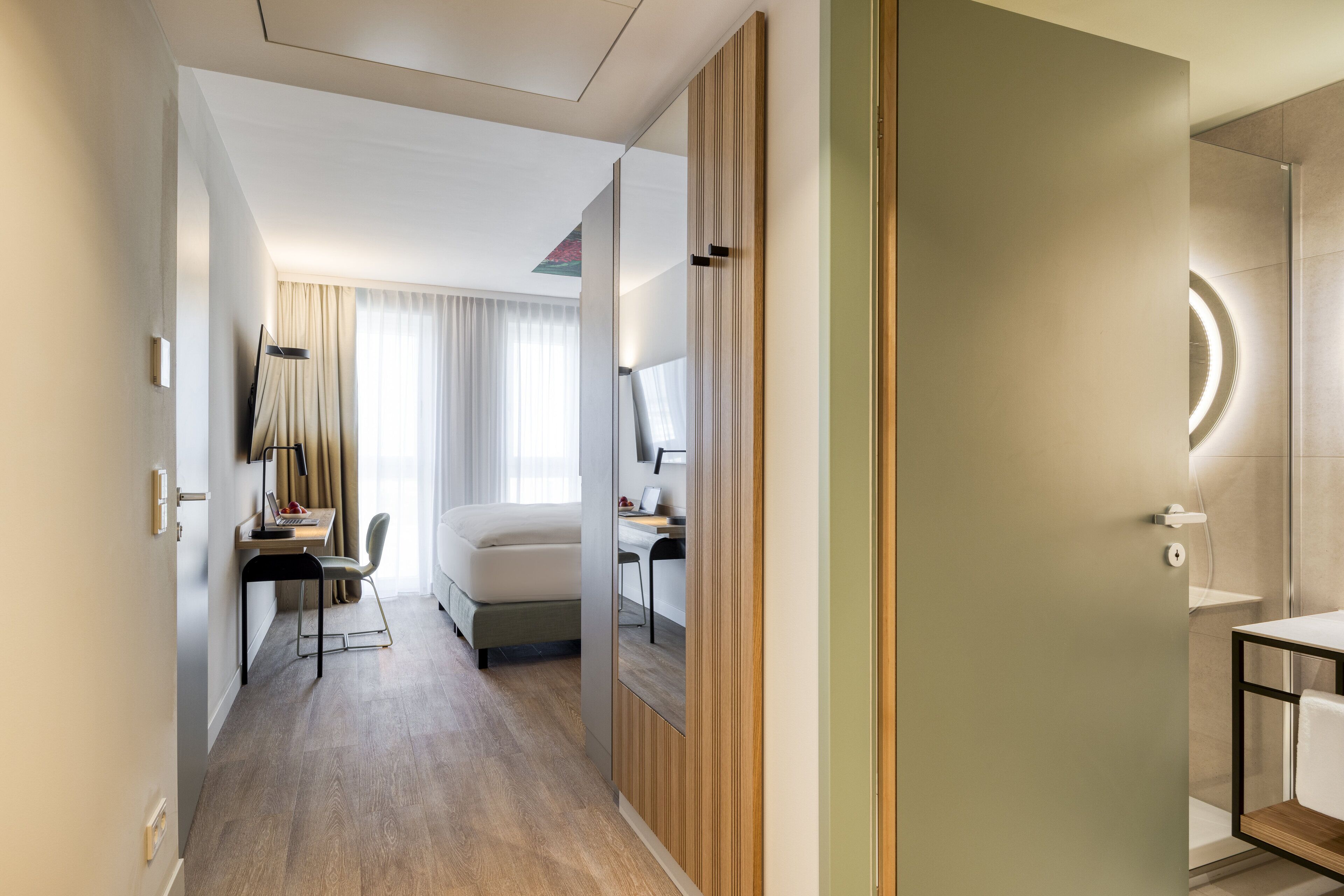A hotel room with a bed, a desk with a chair, a TV, and a bathroom with a mirror and sink.