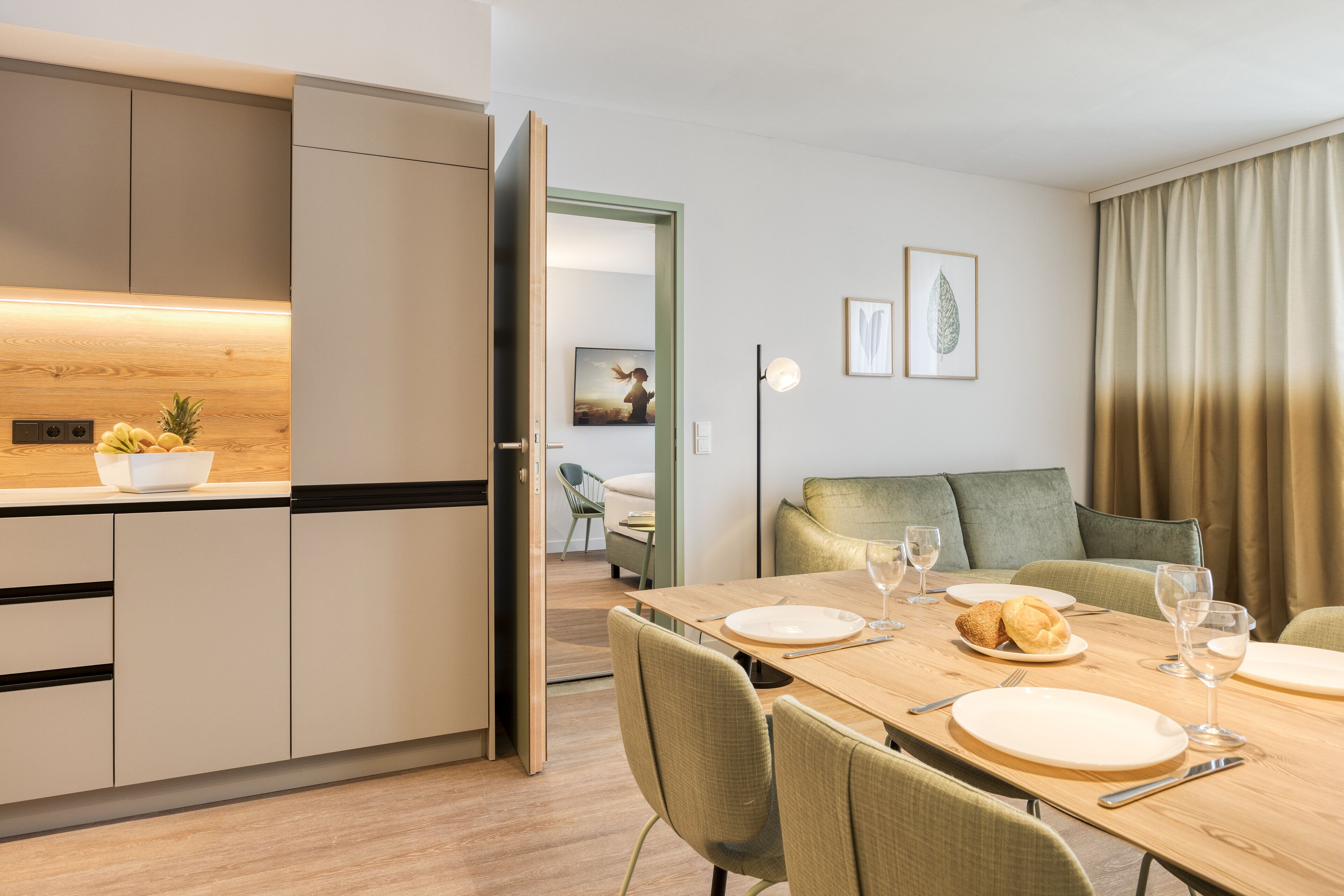 Une cuisine moderne avec un plan de travail en bois, un coin repas avec une table dressée pour quatre personnes, et un salon avec un canapé et un tableau accroché au mur.