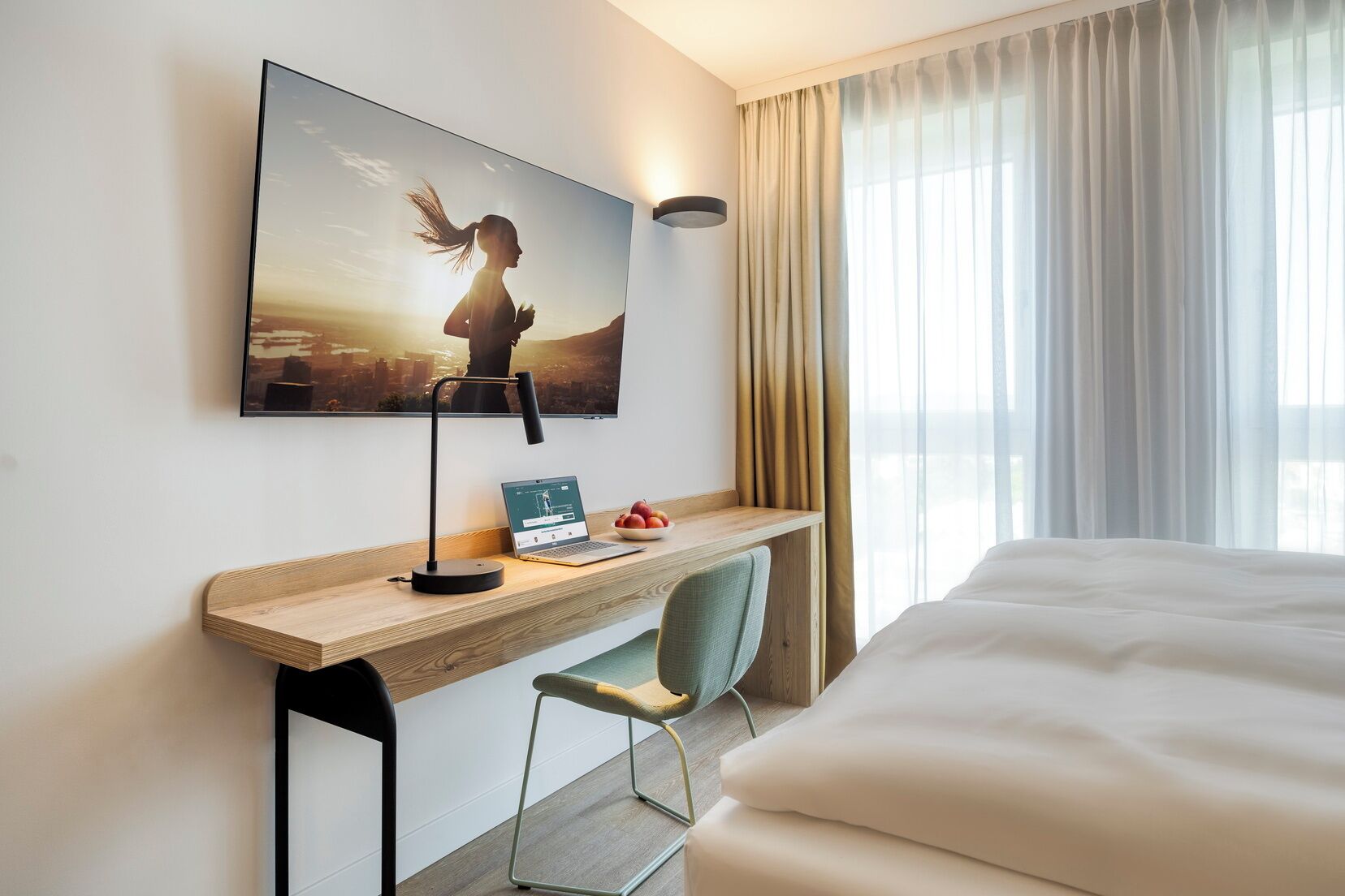 Ein Hotelzimmer mit Bett, Schreibtisch mit Laptop, Stuhl, Wandfernseher und einem Fenster mit Vorhängen.