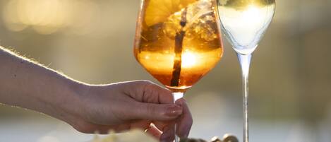 A hand holding a glass with a drink and a straw, and a glass of white wine on a table with a bowl of olives and a small dish.
