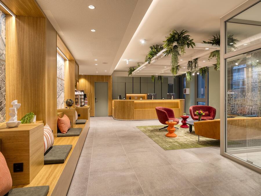 Un área de recepción moderna con un mostrador de madera, asientos con cojines, una silla roja y un muro de cristal con vistas a la ciudad.